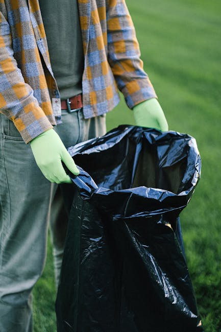 A person standing outdoors on a grassy area, wearing a plaid shirt over a green t-shirt and grey trousers, is holding open a black garbage bag lined with a blue interior. The individual is wearing lig