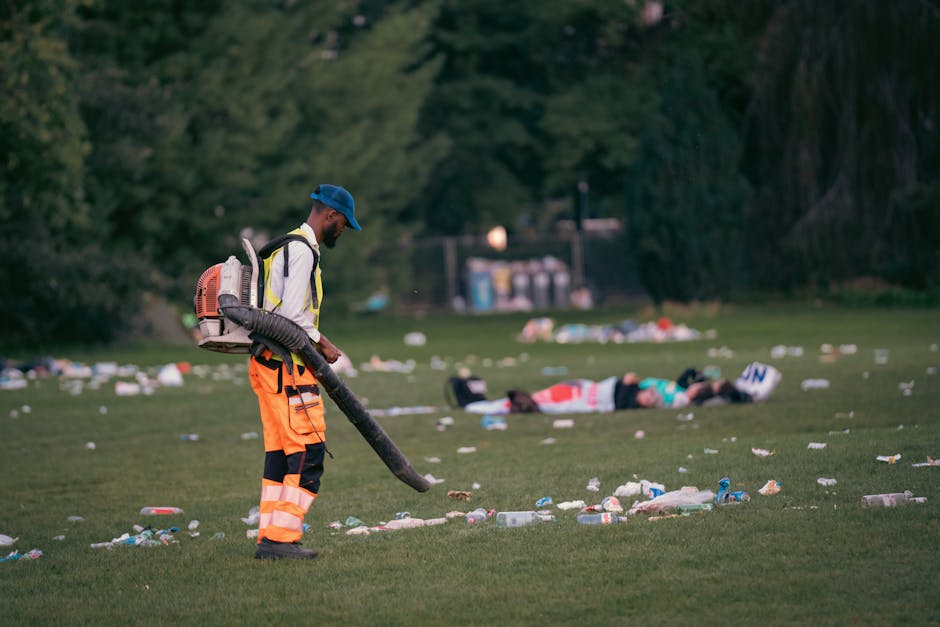 A large, industrial green waste disposal bin positioned outdoors on a city street, filled with various types of rubbish including cardboard boxes, some flattened and others still partially assembled, 